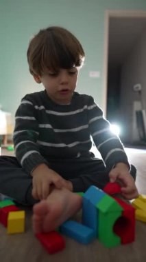 Yatak odasında yapı taşlarıyla oynayan küçük bir çocuk. Evde oturan mutlu çocuk, Dikey Video.mov 'da renkli blok inşaatı ile oynuyor.