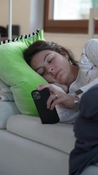 携帯電話の画面をソファの上に寝そべっている退屈な女性 落ち込んでいる人は垂直ビデオでオンラインでソーシャルメディアを閲覧 — ストック動画
