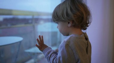 Bored little toddler child wanting to outside stuck at home. Baby standing by window looking out