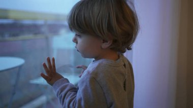 Bored little toddler child wanting to outside stuck at home. Baby standing by window looking out