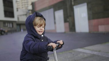 Happy toddler boy riding scooter outside on sidewalk, active child rides scooter