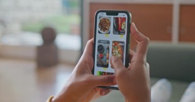 Person browsing restaurant menu online from home. Close up female hand scrolling food delivery app seated at sofa in living room. Comfortable person ordering from smartphone