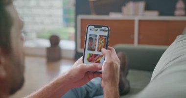 Man browsing restaurant menu online from home. Close up hand scrolling food delivery app seated at sofa in living room. Comfortable person ordering from smartphone