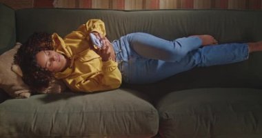 A Brazilian young woman typing message on smartphone device lying on couch at night. A black female person using phone relaxing in sofa smiling. Top view