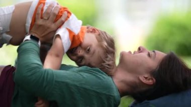 Mother lifting child son in air laying on grass at park outside. Family lifestyle enjoying weekend activity