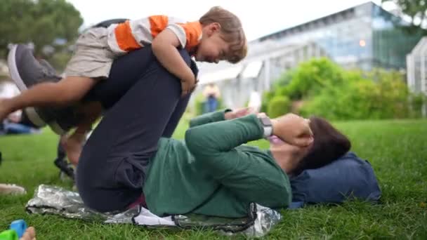 公園の草の上に芝生の上に寝そべっている男の子の息子と遊ぶ遊び心のある母親 親の足で飛ぶ子供 — ストック動画