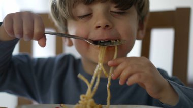 One small child eating pasta noodles by himself with fork. Kid eats lunch meal