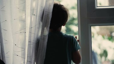 Melankolik küçük çocuk pencerenin kenarında dikilip pencereden dışarı bakıyor.