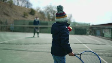 Çocukların kış sezonunda ebeveynleriyle tenis oynamaları. Çocuk palto giyiyor ve bereyle topa vuruyor. Küçük çocuk dışarıda spor yapıyor.