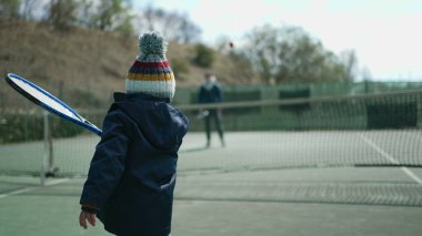 Çocukların kış sezonunda ebeveynleriyle tenis oynamaları. Çocuk palto giyiyor ve bereyle topa vuruyor. Küçük çocuk dışarıda spor yapıyor.