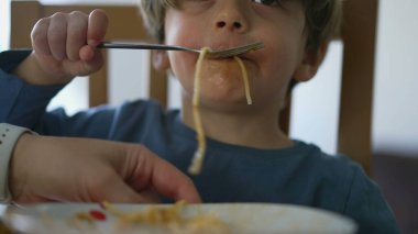 Yakından çocuk suratlı makarna yiyor. Bir sarışın çocuklara makarna yediriyor.