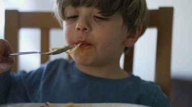 Yakından çocuk suratlı makarna yiyor. Bir sarışın çocuklara makarna yediriyor.
