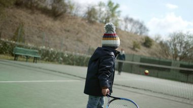 Çocuk tenis raketiyle topa vuruyor kış mevsiminde kışlık kıyafetler giyiyor. Çocuk spor yapıyor.