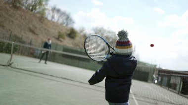 Çocuk tenis raketiyle topa vuruyor kış mevsiminde kışlık kıyafetler giyiyor. Çocuk spor yapıyor.