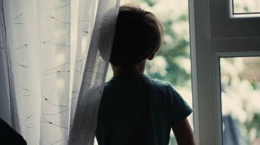 Melankolik küçük çocuk pencerenin kenarında dikilip pencereden dışarı bakıyor.