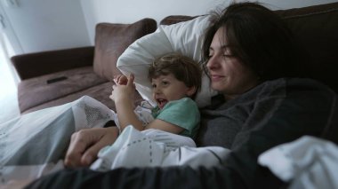 Anne ve çocuk evdeki kanepede yatıyorlar. Anne oğlunu öpüyor, sevgi ve şefkat gösteriyor. Ebeveyn ve çocuk koltukta el çırpıyor.