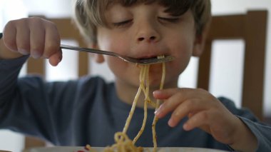 One small child eating pasta noodles by himself with fork. Kid eats lunch meal