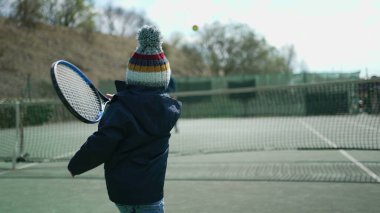 Çocukların kış sezonunda ebeveynleriyle tenis oynamaları. Çocuk palto giyiyor ve bereyle topa vuruyor. Küçük çocuk dışarıda spor yapıyor.