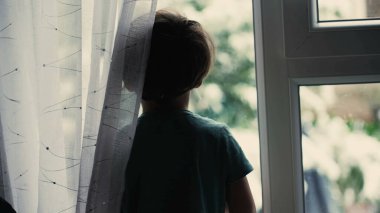 Melankolik küçük çocuk pencerenin kenarında dikilip pencereden dışarı bakıyor.
