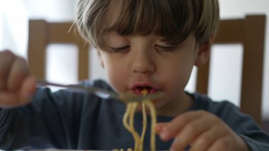 One small child eating pasta noodles by himself with fork. Kid eats lunch meal