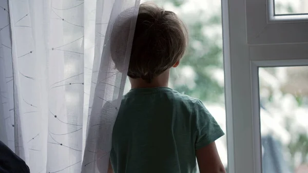 Child standing by window during snowy day. Little boy stands by glass looking outside during winter season