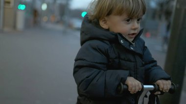 Little toddler boy riding three wheeled scooter in city at night during winter