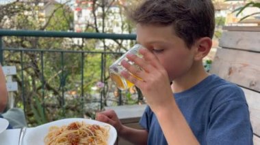 Young boy at lunch table with family outside in home patio. Child eating pasta and drinking juice