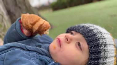 Happy child eating croissant bread laying outside at park. Little boy wearing winter clothes jacket and beanie. Kid snacking carb food.