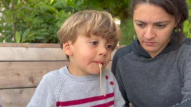 Mother feeding little boy pasta spaghetti outdoors. Child eats and drink at lunch table during sunny day.