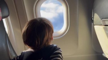 Child traveling by plane. Little boy passenger looking at blue sky pass by through airplane window. Vacation concept