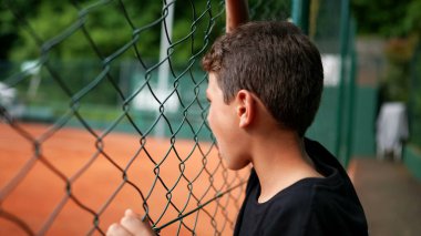 Çocuk çitlere yaslanarak tenis maçı izliyor.