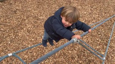 One little baby boy climbing playground structure role outside during winter season. Effortful child playing outdoors wearing jacket climbs upwards