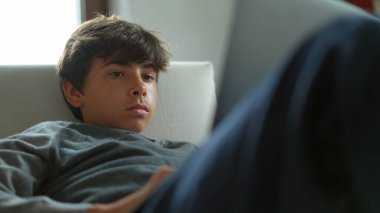 Genç çocuk evde dizüstü bilgisayar kullanıyor. Çocuk koltukta bilgisayar ekranına bakıyor.