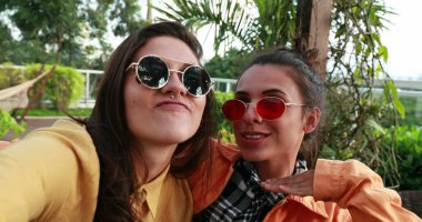 Girlfriends taking selfie posing to camera doing funny faces wearing round sunglasses