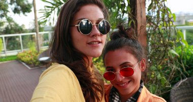 Millennial girls posing for selfie outside. Two young women wearing round sunglasses taking photo smiling