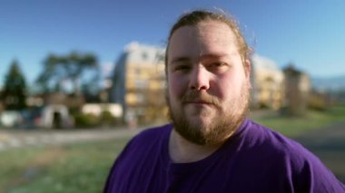 Portrait of a happy overweight young man standing outside looking at camera. One casual male person with beard smiling