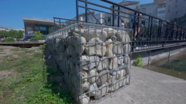 Many rocks stacked behind wire cage outside near bridge. Gray granite stone behind bars. Metal mesh with stones