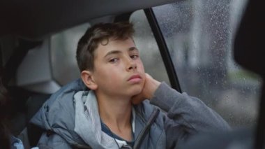 One contemplative young boy in car backseat looking out window during rainy day. Pensive teen kid travels on road