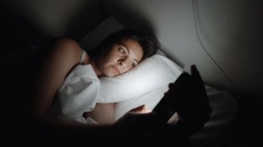 Woman looking at cellphone screen in the dark. Female caucasian person with glowing light coming from smartphone laying in bed at night