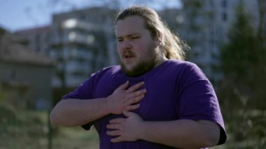 One young overweight man having chest pains standing outdoors. A fat person feeling stressful pain with difficulty in breathing