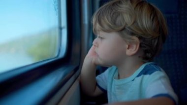 Pensive child travels by train holding chin with hand. Contemplative little boy staring at transportation window