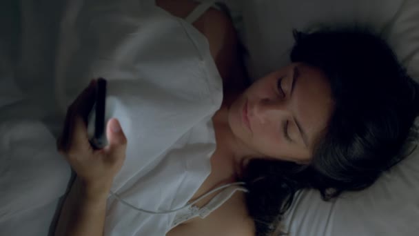女性は夜のチェックスマートフォンでベッドに横たわっていた 寝る前に電話画面を見つめている女性のトップビューの視点 — ストック動画