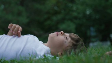 Rahat çocuk parkta çimlere uzandı. Tasasız küçük çocuk doğanın dışında yatıyor. Mutlu çocuk dinleniyor.
