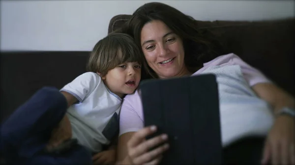 Mother and child looking at tablet device screen together on couch at home. Lifestyle Parent and son staring at technological screen talking with family member