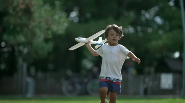 Young boy running with plane toy at park. Carefree Child throwing airplane glider outdoors having fun. Playful kid throws foam plane in sunny beautiful day