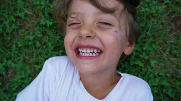 Happy child laughing close up face. Male kid laid on grass smiling. Real life laugh and smile. Authentic joy expression