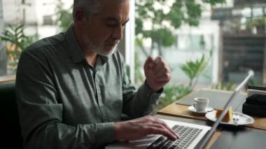 Focused male businessman typing on laptop computer seating at coffee shop. Middle aged executives types email