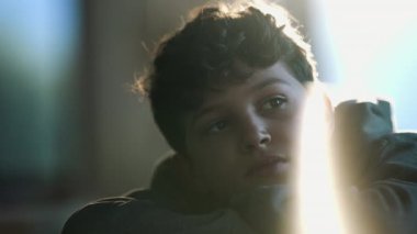 One contemplative kid daydreaming close up face. Pensive preteen kid leaning on table with arms crossed thinking with lens flare artistic reflection
