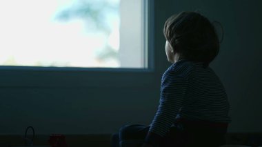 Karanlıkta oturan yalnız küçük bir çocuk pencereye bakıyor. Üzgün çocuk yatak odasında yalnız.