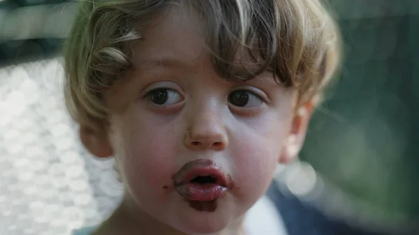 Adorable child boy with dirty mouth covered with chocolate ice cream. Messy kid outside. Contemplative pondering question